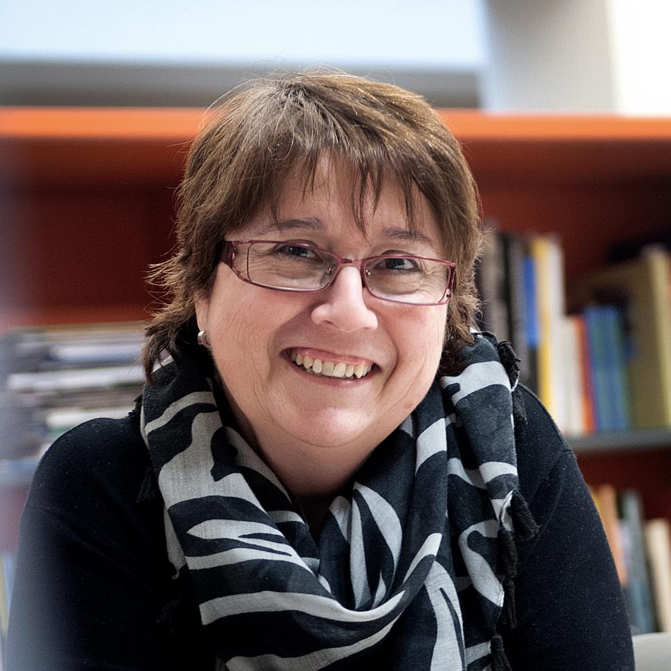 Una mujer sonriente con cabello corto y marrón, usando gafas y un elegante atuendo negro, se encuentra frente a un fondo de estanterías llenas de libros. Lleva una bufanda de patrón negro y blanco, irradiando calidez y profesionalismo.