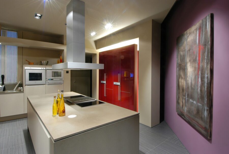 Un interior de cocina contemporánea que presenta una gran isla con un elegante encimera y una estufa integrada, gabinetes de color claro, electrodomésticos empotrados, una striking campana extractora plateada y puertas de refrigerador rojas y audaces, todo sobre paredes neutrales adornadas con obras de arte abstractas. El área se ilumina con luces empotradas y luz natural, con un suelo de azulejos gris claro, ejemplificando la estética del diseño moderno.