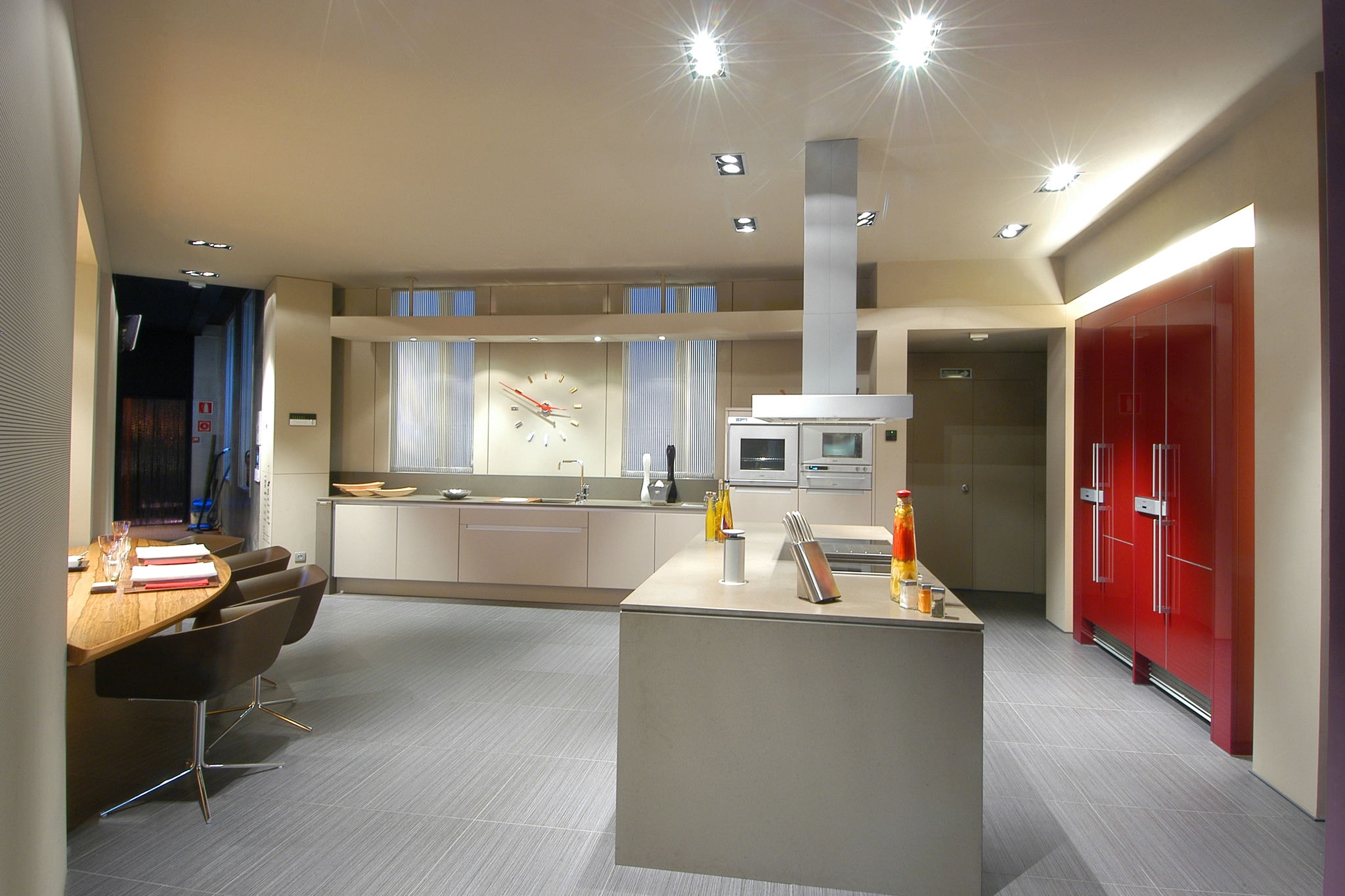 Una cocina contemporánea que muestra una estética elegante y minimalista con una isla central gris, electrodomésticos empotrados y llamativos refrigeradores rojos contra muebles de tonos neutros.
