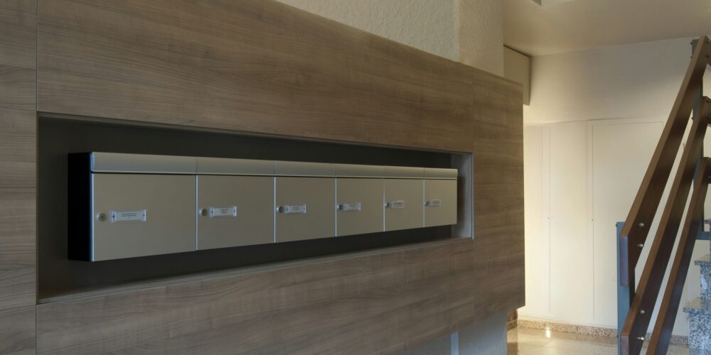 Diseño de interiores contemporáneo en Barcelona que presenta una elegante unidad de buzones montada en la pared sobre un panel de madera, destacando la funcionalidad y materiales de alta calidad.