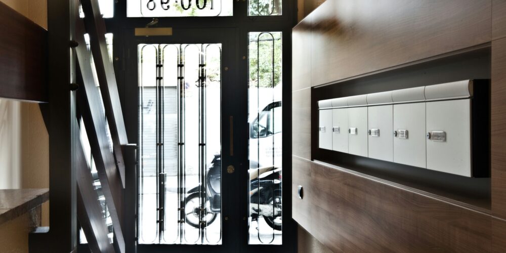 Interiores de un vestíbulo moderno en Barcelona que muestra una puerta de cristal con rejas, luz natural, escaleras contemporáneas y un panel de madera con buzones blancos. Destacan los materiales de alta calidad y los detalles arquitectónicos que reflejan la arquitectura de alta gama y los estilos modernos en el diseño de interiores.