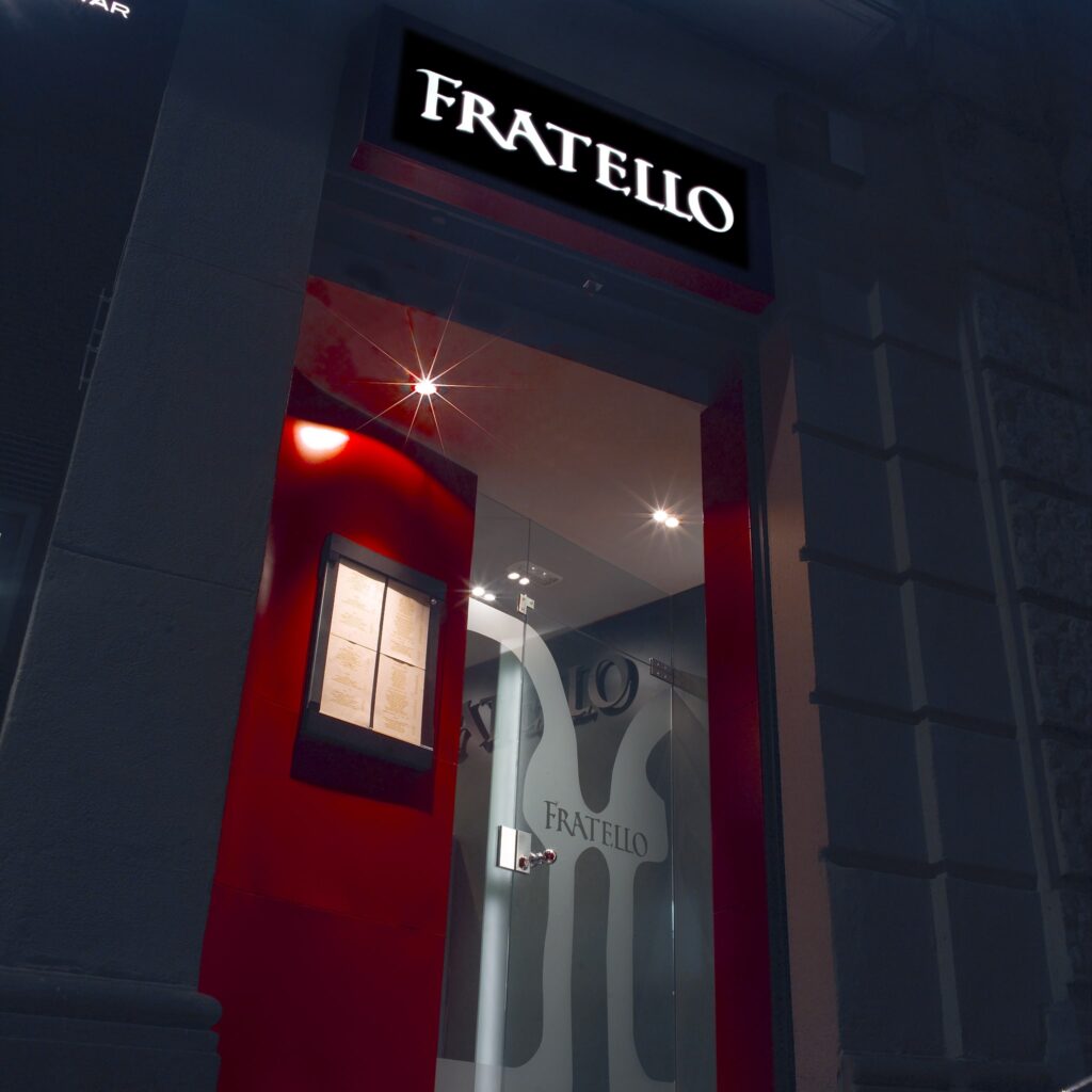 Entrada del Restaurante FRATELLO – Cartel Iluminado y Ambiance Vibrante La entrada del restaurante FRATELLO con letras iluminadas sobre puertas de vidrio, rodeada de una vibrante pared roja y un display del menú, con una luz suave que crea una atmósfera acogedora.