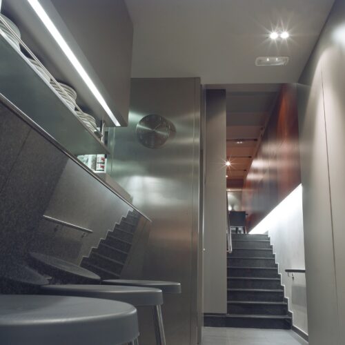 Vista interior de un elegante pasillo moderno con superficies pulidas, mostrador de acero inoxidable y decoración minimalista, iluminado suavemente con escaleras iluminadas y un reloj de pared.