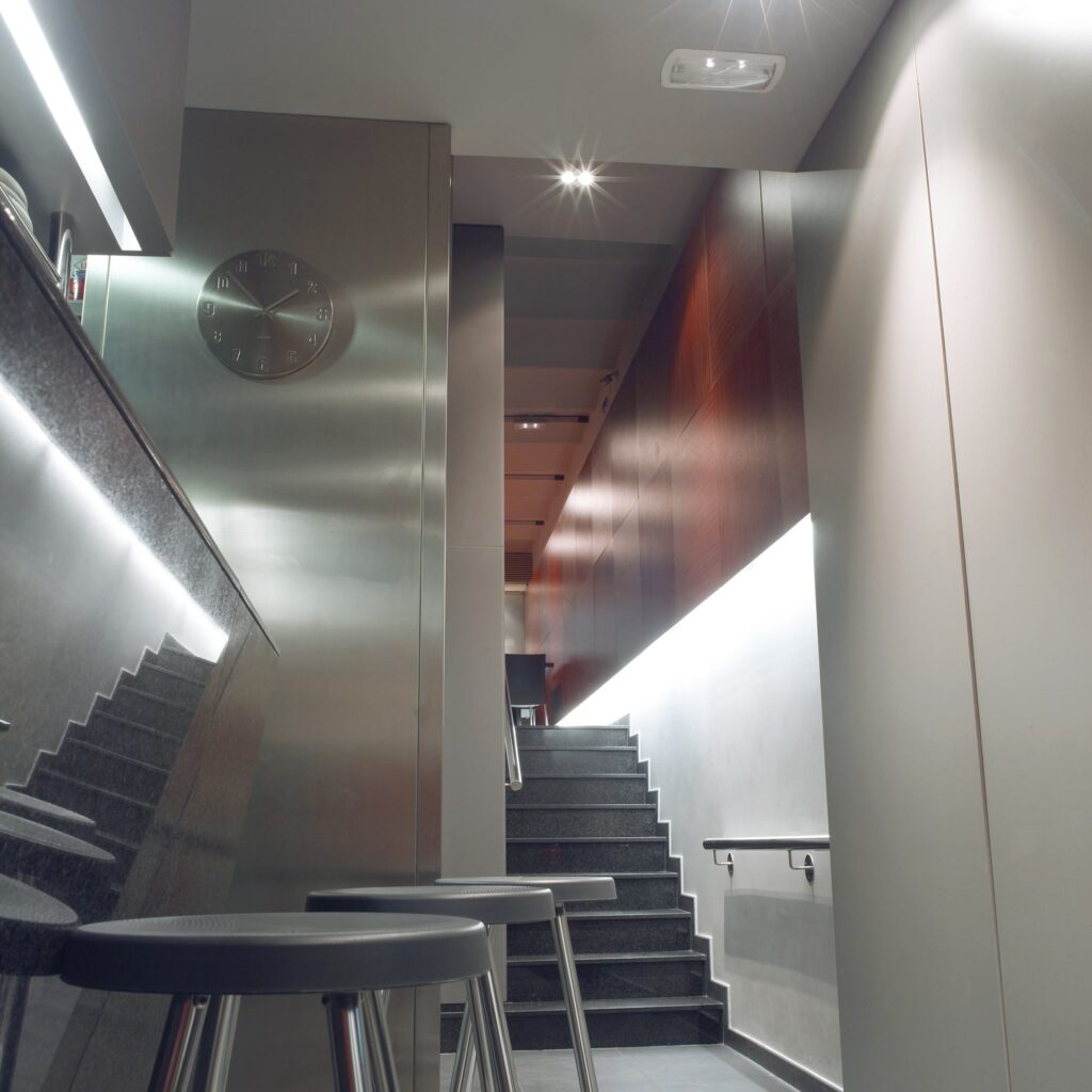 Pasillo Moderno Sofisticado con Elementos de Diseño Minimalista Un pasillo moderno bien iluminado que conduce a una escalera, con un diseño minimalista en tonos grises y marrones, taburetes simples y una iluminación en las paredes que acentúa las texturas.
