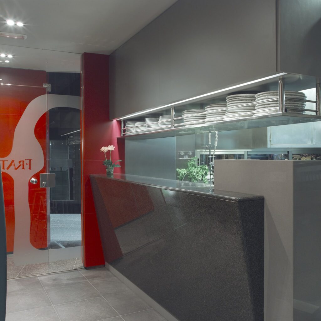 Interiores de Restaurante Moderno y Elegante con Mostrador de Granito Negro Un interior de restaurante contemporáneo que muestra un mostrador de granito negro pulido con una estantería de acero inoxidable encima, exhibiendo platos blancos apilados ordenadamente. El fondo presenta paredes pintadas de gris y rojo, complementadas por una pequeña planta en maceta, creando una atmósfera acogedora. Una puerta de vidrio revela parcialmente la imagen de marca del restaurante.
