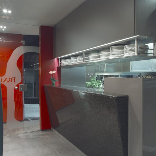 Un interior de restaurante contemporáneo que muestra un mostrador de granito negro pulido con una estantería de acero inoxidable encima, exhibiendo platos blancos apilados ordenadamente. El fondo presenta paredes pintadas de gris y rojo, complementadas por una pequeña planta en maceta, creando una atmósfera acogedora. Una puerta de vidrio revela parcialmente la imagen de marca del restaurante.