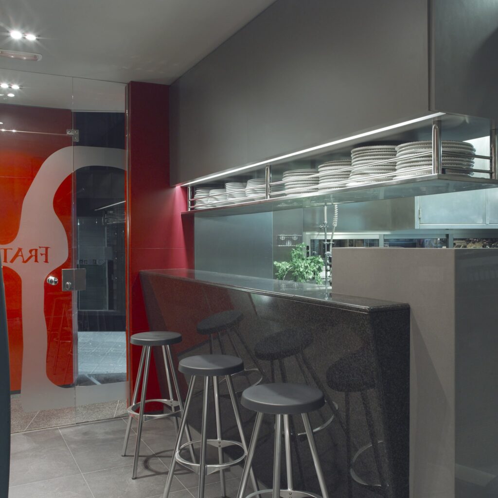 Interiores de Restaurante Contemporáneo con Barra de Granito Negro y Ambiente Acogedor Un restaurante moderno que muestra una elegante barra de granito negro combinada con taburetes de bar de metal con estilo. El interior cuenta con una estantería bien iluminada adornada con platos blancos apilados ordenadamente, acentuada por paredes en sutiles tonos grises y rojos. Una puerta de vidrio resalta la marca del restaurante, creando una atmósfera acogedora perfecta para cenar y socializar.
