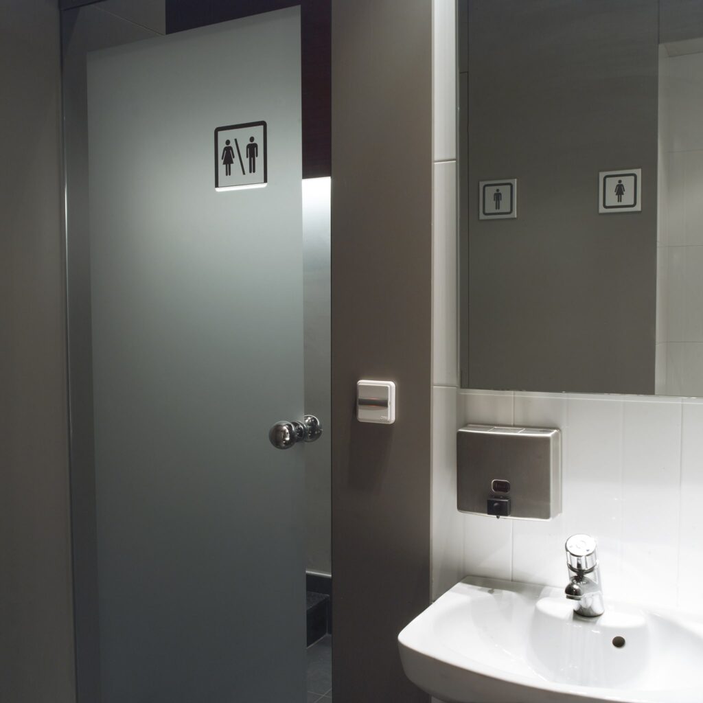 Interior de Baño Unisex Moderno con Puerta de Cristal Esmerilado Un baño moderno que presenta una puerta de cristal esmerilado con símbolos de masculino y femenino, mostrando un diseño elegante con un lavabo blanco, grifo moderno, secador de manos y un gran espejo, todo en tonos de gris y blanco.