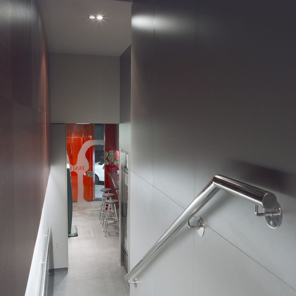 Pasillo moderno con barandilla de acero inoxidable y puerta de vidrio Un pasillo interior contemporáneo que presenta una elegante barandilla de acero inoxidable, paredes de panel oscuro que contrastan con los azulejos de piso gris claro, y una puerta de vidrio que conduce a una zona temática en rojo con taburetes de barra, creando una atmósfera limpia y moderna.