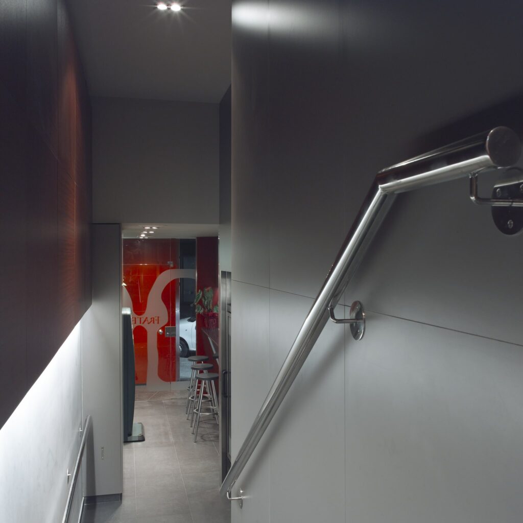 Escalera interior contemporánea con pasamanos de metal y partición de vidrio Una escalera interior moderna que presenta un elegante pasamanos de metal y paredes grises iluminadas por una suave luz artificial. En la parte superior, una partición de vidrio muestra un audaz logo rojo, con taburetes de bar y muebles visibles que crean una atmósfera vibrante.