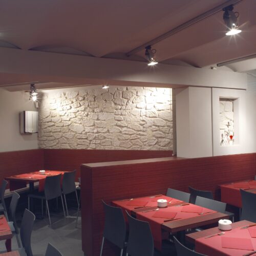 Interior de un restaurante moderno que presenta mesas de comedor acogedoras con manteles rojos, cubiertos y servilleteros blancos, paredes de piedra expuesta, focos cálidos montados en el techo, y un panel de madera para privacidad.