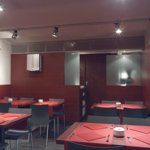 Vista interior de un restaurante contemporáneo que muestra mesas adornadas con manteles rojos, sillas minimalistas y una iluminación suave en riel que realza la atmósfera cálida. Las paredes con acabado de madera rica y los paneles de vidrio esmerilado añaden elegancia, con una visión parcial del área de la cocina visible a través del vidrio.
