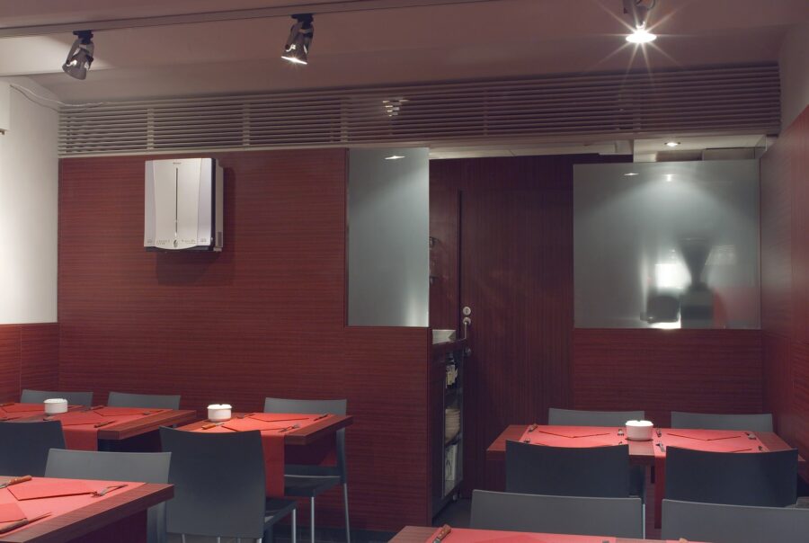 Vista interior de un restaurante contemporáneo que muestra mesas adornadas con manteles rojos, sillas minimalistas y una iluminación suave en riel que realza la atmósfera cálida. Las paredes con acabado de madera rica y los paneles de vidrio esmerilado añaden elegancia, con una visión parcial del área de la cocina visible a través del vidrio.