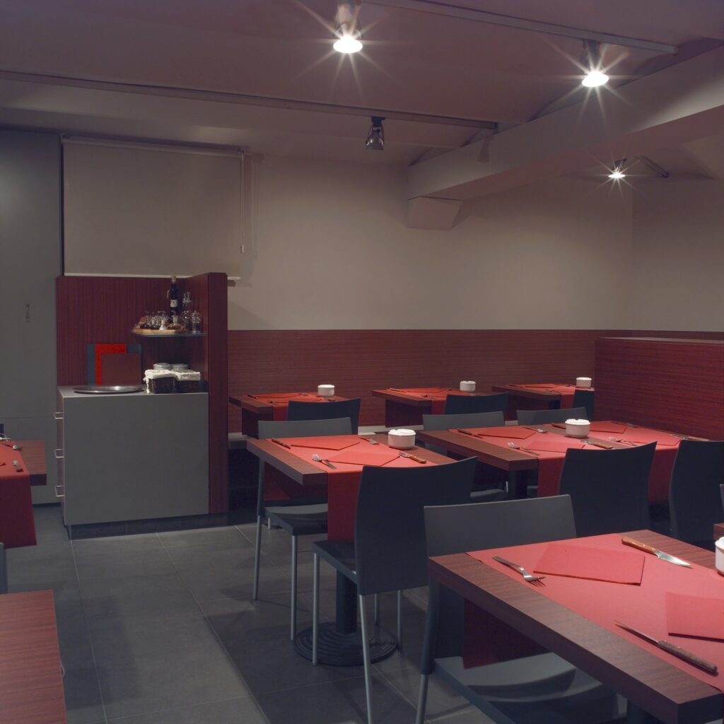Interiores de Restaurante Moderno Minimalista con Iluminación Cálida y Decoración Elegante Comedores: Interior de un restaurante contemporáneo minimalista que muestra suelos de madera oscura, iluminación ambiental cálida, sillas negras elegantes y mesas adornadas con servilletas rojas y vajilla blanca, creando una atmósfera acogedora para una experiencia gastronómica casual pero sofisticada.