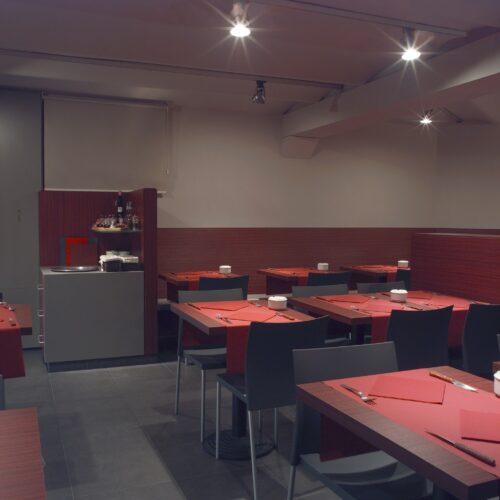 Comedores: Interior de un restaurante contemporáneo minimalista que muestra suelos de madera oscura, iluminación ambiental cálida, sillas negras elegantes y mesas adornadas con servilletas rojas y vajilla blanca, creando una atmósfera acogedora para una experiencia gastronómica casual pero sofisticada.
