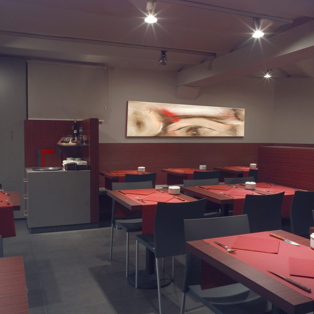 Diseño de Interior de Restaurante Moderno y Elegante con Ambiente Acogedor Interior de un restaurante contemporáneo que presenta mesas de comedor de madera con servilletas rojas y cubiertos, sillas negras, un mostrador gris que exhibe botellas y tarros, y arte abstracto en la pared. La iluminación cálida crea una atmósfera acogedora para los comensales.