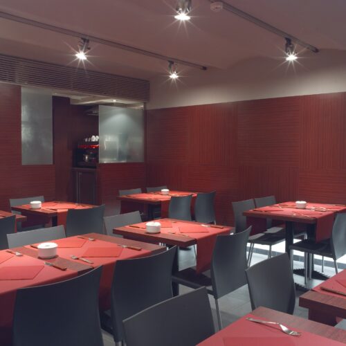 Pizzería Fratello: Un interior de restaurante moderno que presenta mesas ordenadamente dispuestas, cada una con manteles rojos y utensilios, paredes marrones cálidas y luces brillantes en el techo, mostrando un diseño contemporáneo y una atmósfera acogedora.