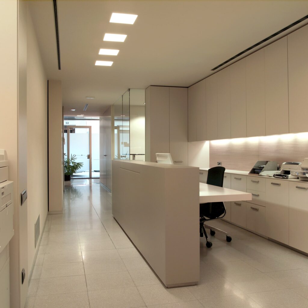 Área de recepción de oficina moderna con diseño elegante Área de recepción de oficina moderna, bien iluminada, que presenta un diseño minimalista con un mostrador de recepción de color claro, silla de oficina negra, equipo de oficina, suelo de baldosas pulidas y un toque de vegetación de una planta cercana.