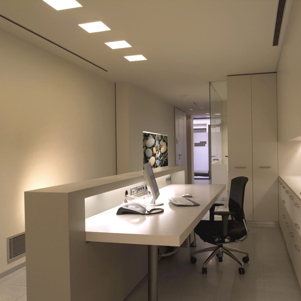 Espacio de Oficina Moderno y Minimalista con Diseño Ergonómico Oficina moderna bien iluminada que presenta un escritorio blanco elegante con equipo informático, una silla ergonómica negra y arte mural de acento que muestra piedras lisas, complementado por altos armarios blancos y una partición de vidrio.
