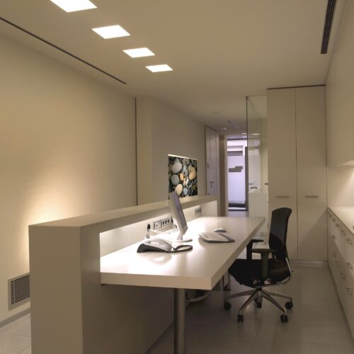 Oficina moderna bien iluminada que presenta un escritorio blanco elegante con equipo informático, una silla ergonómica negra y arte mural de acento que muestra piedras lisas, complementado por altos armarios blancos y una partición de vidrio.