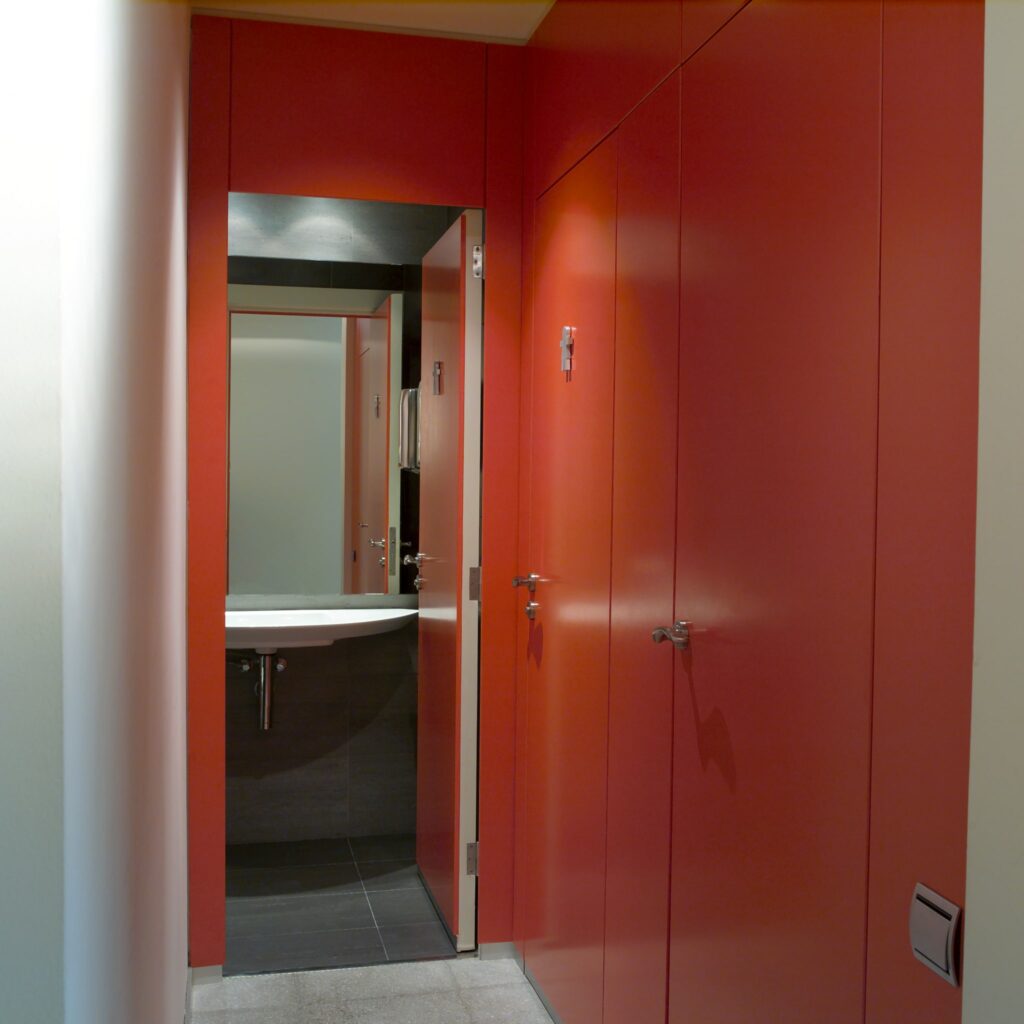 Pasillo Moderno y Vibrante que Lleva al Área de Baños Un pasillo contemporáneo con vibrantes paredes naranjas, que conduce a un área de baños con un lavabo blanco y un suelo de baldosas oscuras. Las líneas limpias y un diseño minimalista crean una atmósfera acogedora.