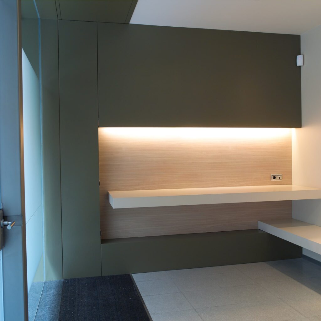 Diseño de Interior Minimalista Contemporáneo con Acentos Verdes Suaves Un interior elegante y moderno que presenta un diseño minimalista. Las paredes están pintadas de un verde suave con un panel de madera acentuado por una iluminación cálida. Una estantería blanca recorre la pared, y grandes baldosas grises cubren el suelo. Una puerta parcialmente visible insinúa un vestíbulo, contribuyendo a la estética contemporánea general.