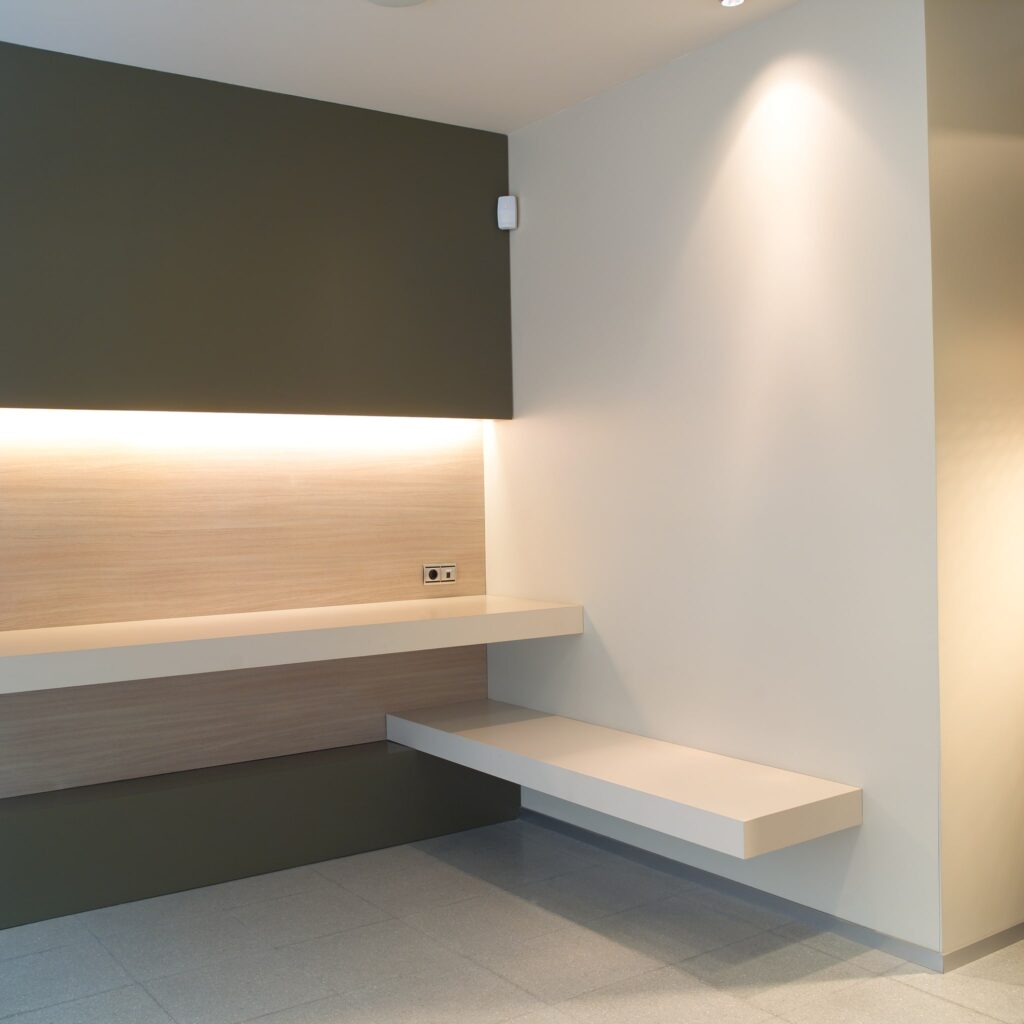 Interior Minimalista Moderno con Estanterías Flotantes y Iluminación Cálida Una escena interior contemporánea que muestra una pared con dos estanterías flotantes: una estantería superior de madera clara y una estantería inferior blanca y elegante, iluminadas por una suave y cálida luz. La pared presenta un color verde apagado que contrasta con el entorno de tonos beige y blanco, enfatizando una estética limpia y funcional.