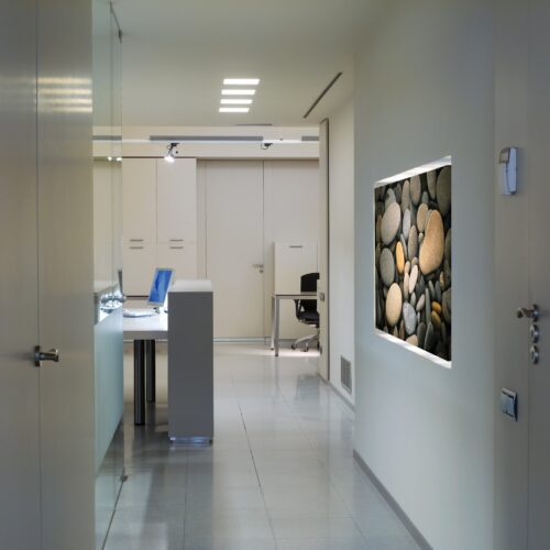 Oficinas Intersa: Un pasillo interior moderno que muestra un diseño limpio y una paleta de colores sutil. El pasillo presenta obras de arte enmarcadas de piedras lisas en la pared derecha y conduce a un espacio de trabajo equipado con un escritorio, silla y laptop. La suave iluminación en el techo ilumina el área, creando una atmósfera acogedora.