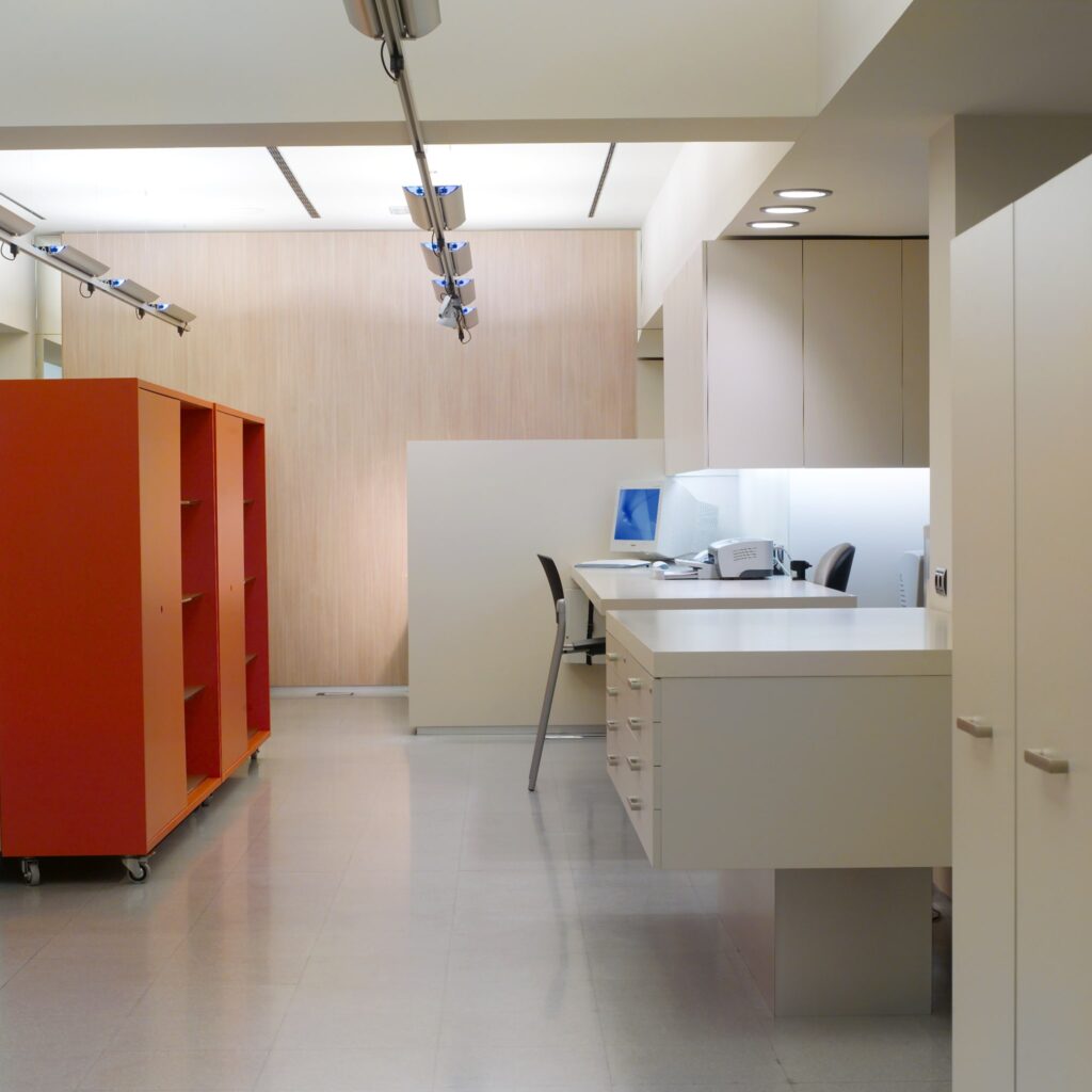 Interior de Oficina Moderno y Minimalista con Iluminación Brillante y Diseño Funcional Imagen de un interior de oficina moderno con un diseño limpio y minimalista, con una pared de color claro, acentos de acabado en madera, área de escritorio blanca equipada con una computadora y suministros de oficina, unidad de estantería naranja con ruedas, piso de baldosas ligeras pulidas, iluminación de riel y una silla negra en el escritorio, creando un espacio de trabajo profesional y organizado.