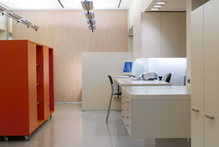 Imagen de un interior de oficina moderno con un diseño limpio y minimalista, con una pared de color claro, acentos de acabado en madera, área de escritorio blanca equipada con una computadora y suministros de oficina, unidad de estantería naranja con ruedas, piso de baldosas ligeras pulidas, iluminación de riel y una silla negra en el escritorio, creando un espacio de trabajo profesional y organizado.