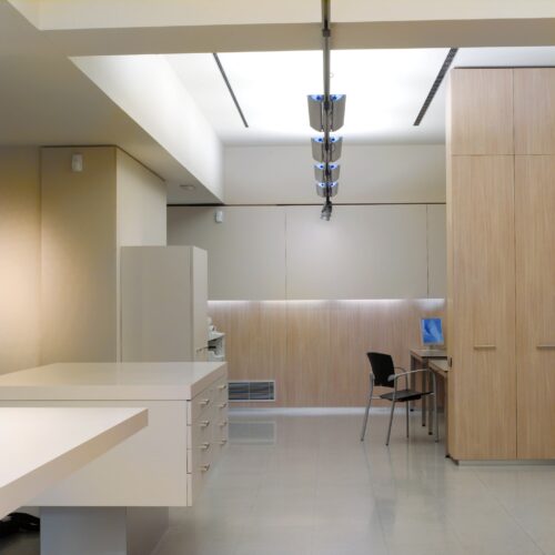 Un interior de oficina moderna que muestra un diseño minimalista con una encimera blanca, unidad de almacenamiento de madera y un elegante escritorio y silla negros. Las paredes son de un beige suave con acentos de paneles de madera, presentando luminarias contemporáneas y un suelo pulido.