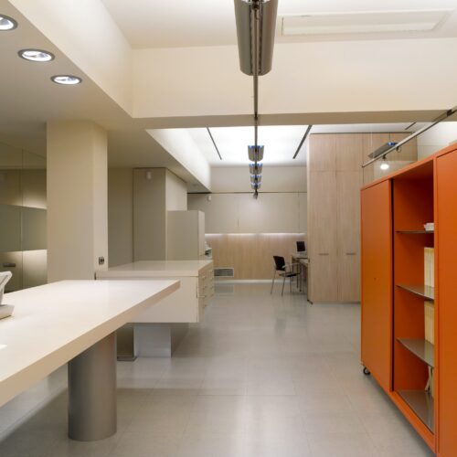 Un interior moderno minimalista y espacioso que presenta líneas limpias y colores neutros, exhibiendo una gran encimera blanca, un suelo de tonos claros, armarios de madera y una unidad de estantería naranja brillante que añade un toque vibrante al espacio de trabajo organizado.