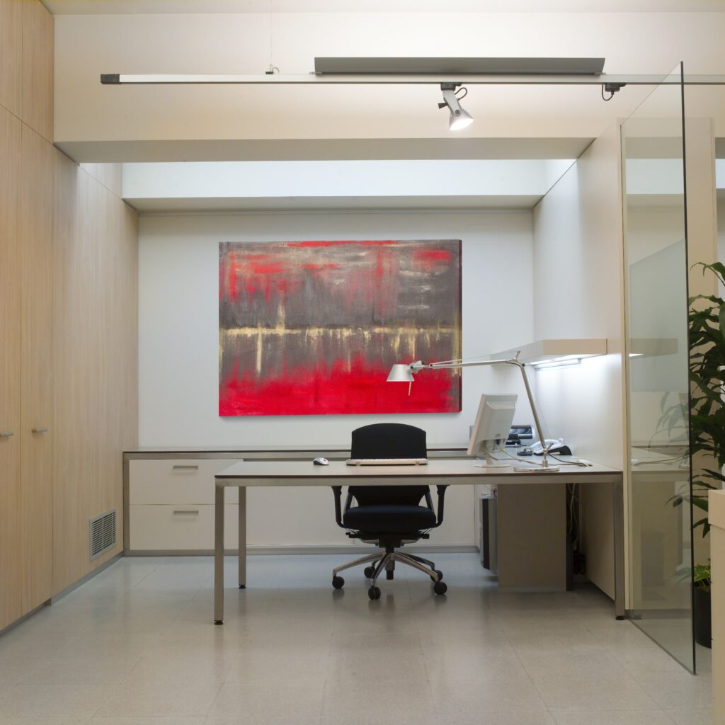 Interior de oficina moderna con un toque artístico Moderna oficina elegante que presenta una silla ergonómica negra, un equipo de computadora en un escritorio estiloso y una pintura vibrante con colores rojos y grises colgando arriba. La oficina tiene gabinetes de madera clara, una pared de vidrio para luz natural y una lámpara de escritorio decorativa complementada por una planta verde.