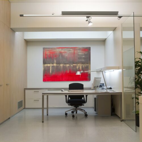 Moderna oficina elegante que presenta una silla ergonómica negra, un equipo de computadora en un escritorio estiloso y una pintura vibrante con colores rojos y grises colgando arriba. La oficina tiene gabinetes de madera clara, una pared de vidrio para luz natural y una lámpara de escritorio decorativa complementada por una planta verde.
