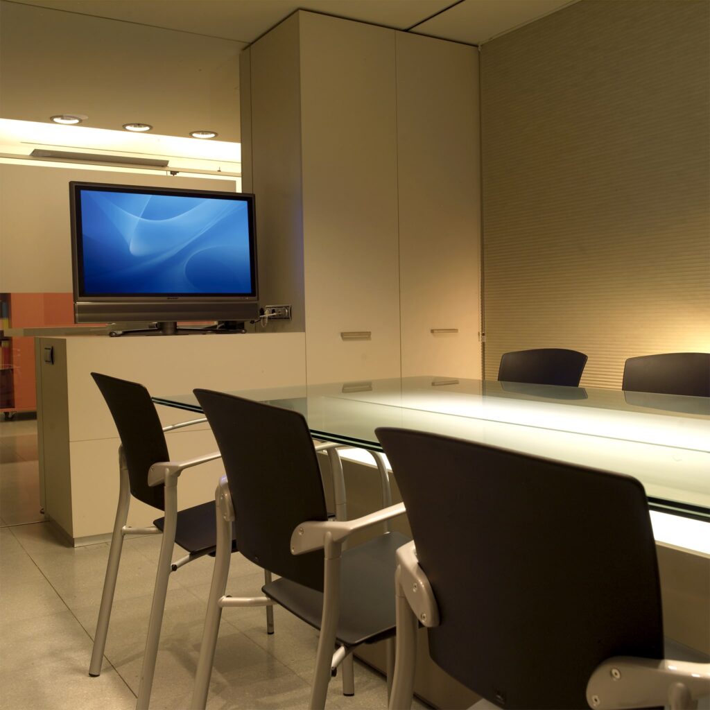 Sala de Conferencias Moderna con Mesa de Vidrio y Televisor de Pantalla Plana Una sala de conferencias moderna y elegante que presenta una mesa de vidrio rodeada de sillas negras, con un televisor de pantalla plana montado en la pared que muestra un fondo azul suave, iluminación debajo del mueble, y paredes texturizadas de color claro, creando una atmósfera profesional adecuada para reuniones.