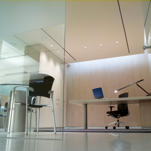 Un interior de oficina contemporáneo que presenta un tabique de vidrio, un escritorio moderno con una computadora de pantalla azul y lámpara de escritorio, una elegante silla de oficina negra y una mesa redonda con una silla negra contra paredes de madera clara, destacando un entorno de trabajo profesional y espacioso.