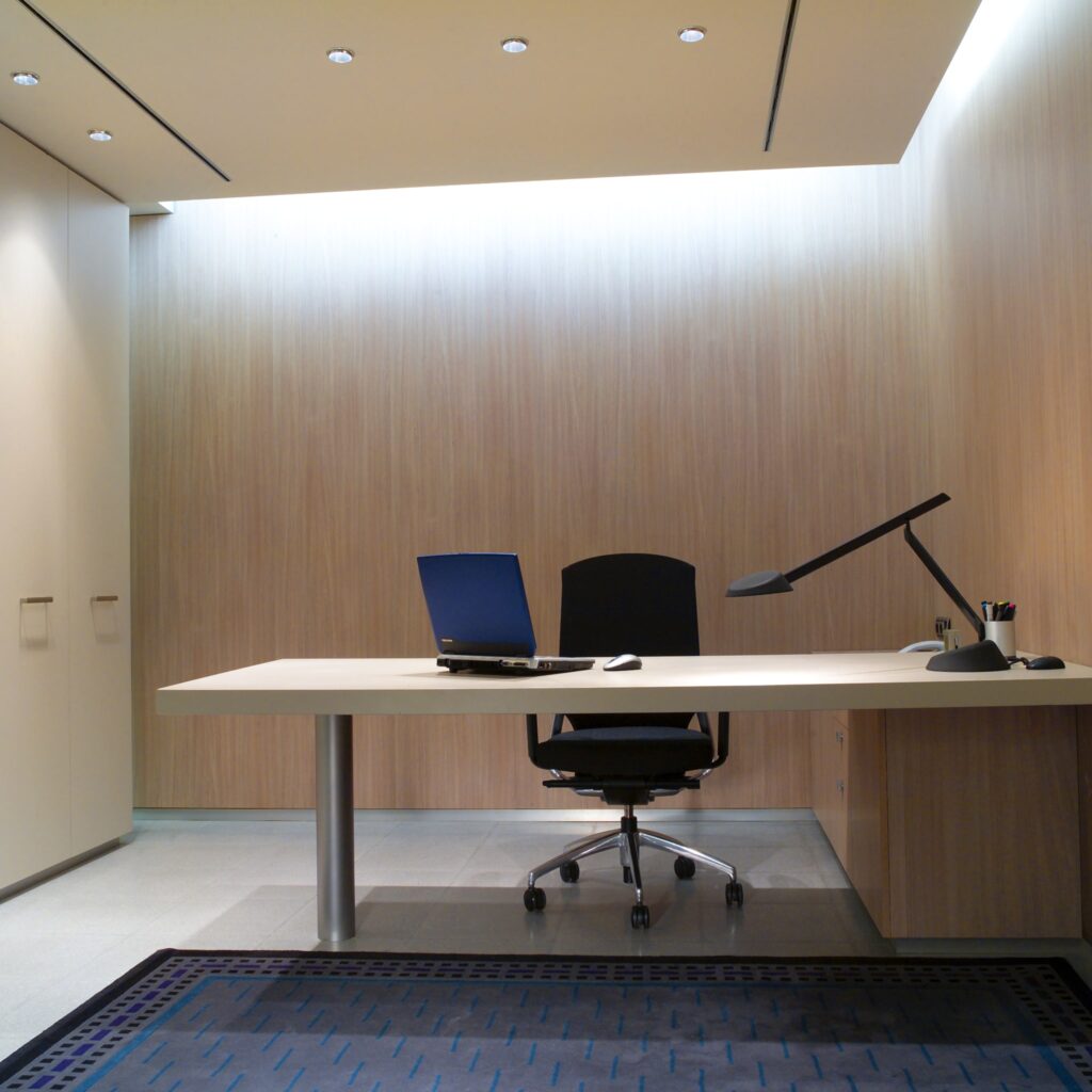 Espacio de Oficina Moderno y Minimalista con Diseño Ergonómico Zonas de trabajo: Un interior de oficina elegante y moderno que presenta una pared de madera minimalista con iluminación ambiental, un escritorio blanco elegante con una laptop, un ratón y una lámpara de escritorio, complementado por una silla ergonómica negra y una alfombra azul con patrón, creando una atmósfera cálida y profesional.
