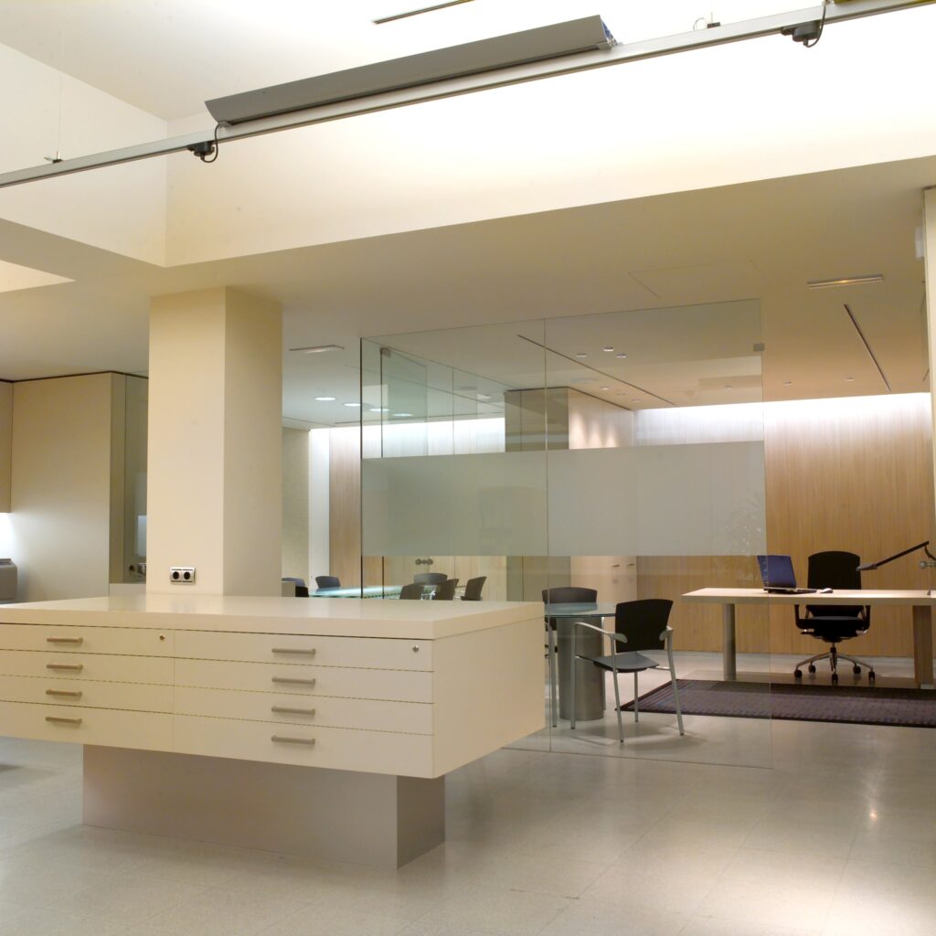 Oficina Moderna y Elegante con Área de Recepción Minimalista Un interior de oficina moderna que muestra un escritorio de recepción blanco y minimalista con múltiples cajones. En el fondo, se puede ver una sala de conferencias con paredes de vidrio, que cuenta con sillas y un escritorio, todo iluminado por una suave luz cenital. Los colores neutros de las paredes crean una atmósfera limpia y profesional.