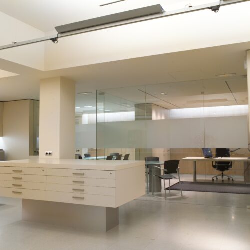 Área de recepción de oficina moderna con un escritorio blanco, partición de vidrio, mesa de conferencias y sillas ergonómicas en un espacio bien iluminado con colores neutros.