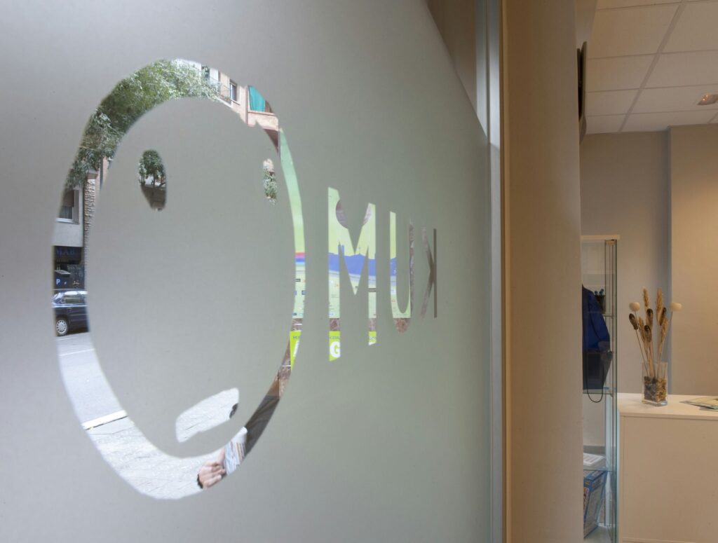 Kumon Barcelona: Ventana de vidrio esmerilado con un logo circular que asemeja un rostro minimalista con las letras 'M' y 'U' en una fuente en negrita. Se puede ver una calle de la ciudad con árboles y coches estacionados al fondo, mostrando un interior moderno decorado con plantas.