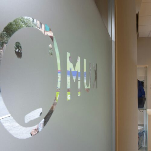 Kumon Barcelona: Ventana de vidrio esmerilado con un logo circular que asemeja un rostro minimalista con las letras 'M' y 'U' en una fuente en negrita. Se puede ver una calle de la ciudad con árboles y coches estacionados al fondo, mostrando un interior moderno decorado con plantas.