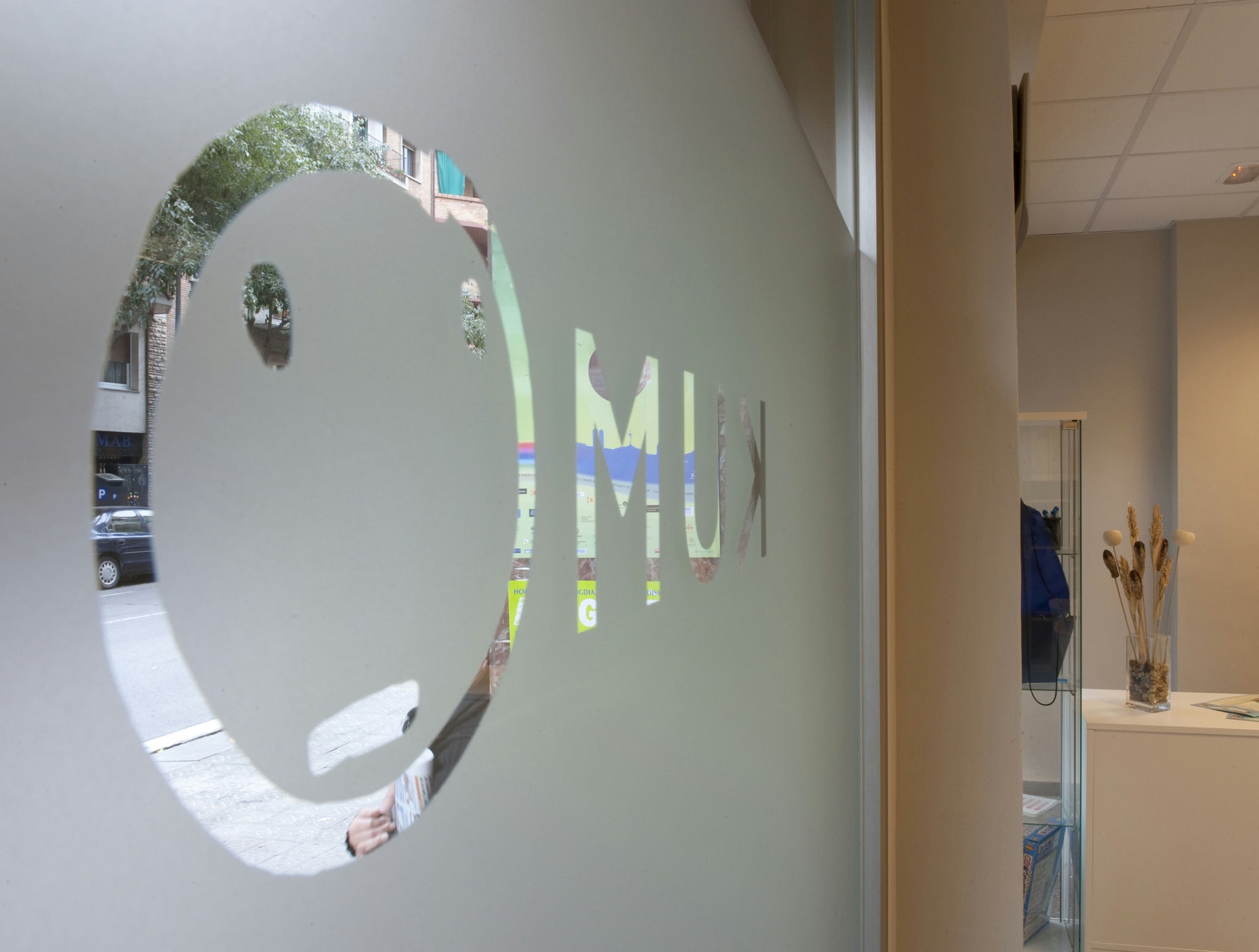 Kumon Barcelona: Ventana de vidrio esmerilado con un logo circular que asemeja un rostro minimalista con las letras 'M' y 'U' en una fuente en negrita. Se puede ver una calle de la ciudad con árboles y coches estacionados al fondo, mostrando un interior moderno decorado con plantas.