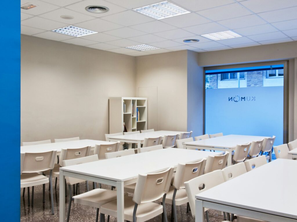 Aula Espaciosa e Inviting para Actividades Educativas Una aula bien iluminada y organizada que cuenta con mesas blancas, paredes de un beige claro, una sección de pared azul y asientos amplios, diseñada para el aprendizaje y los talleres.