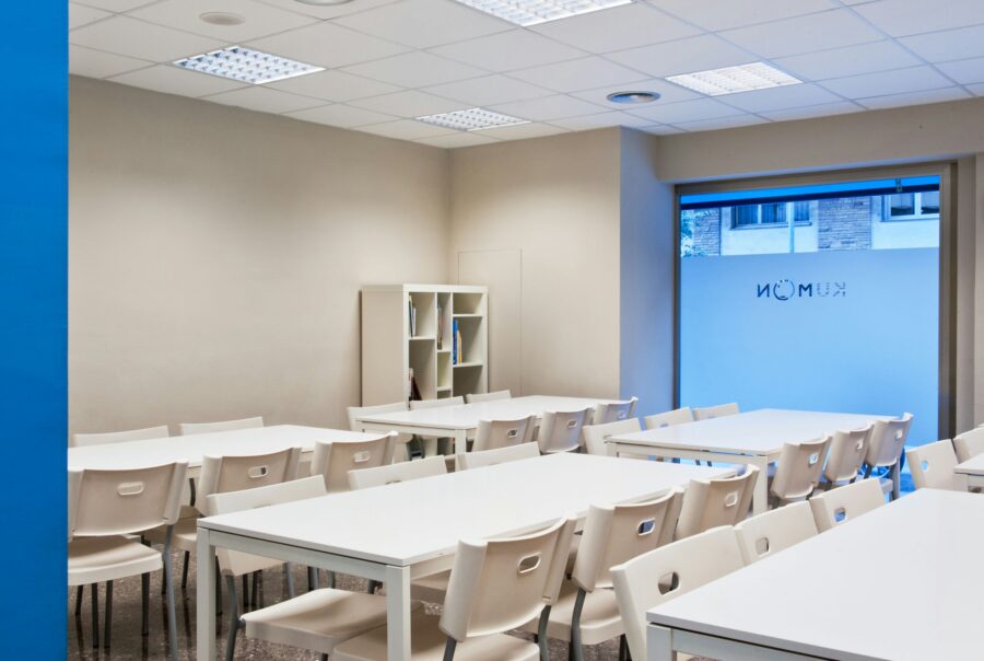 Una aula bien iluminada y organizada que cuenta con mesas blancas, paredes de un beige claro, una sección de pared azul y asientos amplios, diseñada para el aprendizaje y los talleres.
