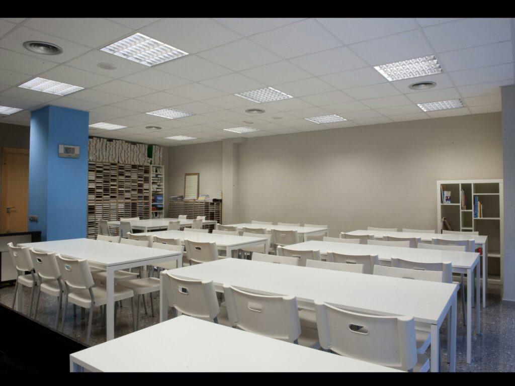 Configuración Moderna del Aula para una Experiencia de Aprendizaje Mejorada Un aula moderna bien iluminada con mesas blancas, sillas a juego y grandes ventanas, con estanterías organizadas llenas de libros y un tabique azul, creando un entorno de estudio propicio.