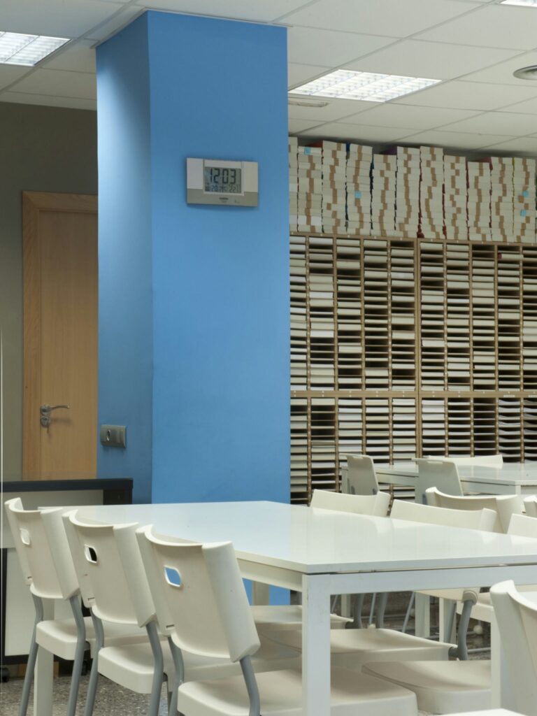 Entorno de Oficina Espacioso con Espacio de Trabajo Organizado Una oficina luminosa y espaciosa con una pared azul claro y un reloj que marca las 12:03, una mesa blanca rodeada de sillas blancas y estantes llenos de archivos apilados ordenadamente, creando una atmósfera organizada y profesional con un suelo gris y luces fluorescentes.