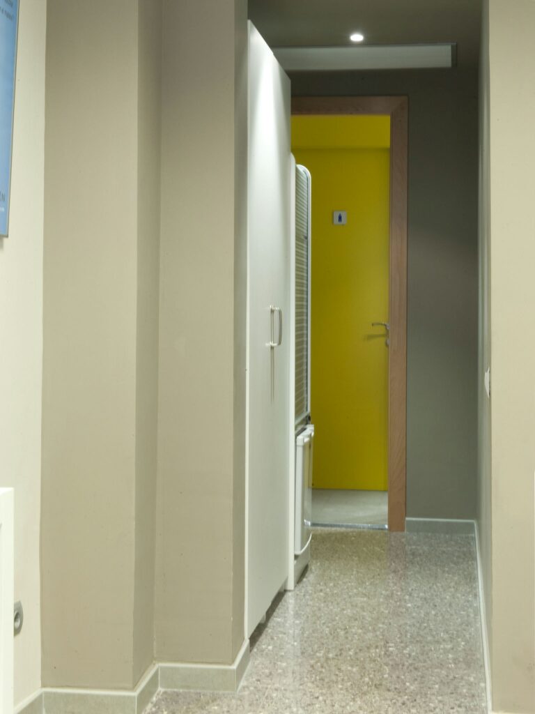 Pasillo moderno que conduce a una puerta amarilla brillante Un pasillo moderno bien iluminado con un suelo de baldosas moteadas que conduce a una puerta amarilla vibrante. En el lado izquierdo, un armario o refrigerador blanco añade al diseño limpio, mientras que las paredes de colores neutros crean una atmósfera serena.