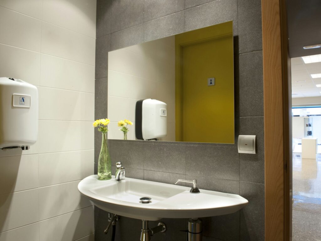 Diseño de Baño Moderno con Detalles Elegantes Un baño moderno y elegante con un lavabo y grifo blancos, paredes de losa gris, y un alto jarrón de vidrio con flores amarillas para una atmósfera fresca. Incluye dispensadores automáticos de jabón y secador de manos, un espejo grande, y una puerta etiquetada en amarillo para uso masculino en el fondo.