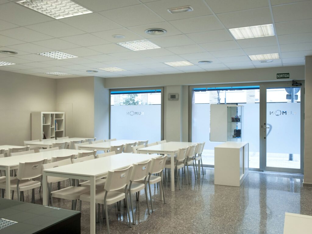 Aula Moderna Espaciosa con Luz Natural Una aula moderna y luminosa y espaciosa que presenta mesas y sillas blancas dispuestas en filas, grandes ventanas que permiten la entrada de luz natural, paredes de color neutro, suelo pulido, una pequeña estantería y un escaparate minimalista cerca de la entrada.