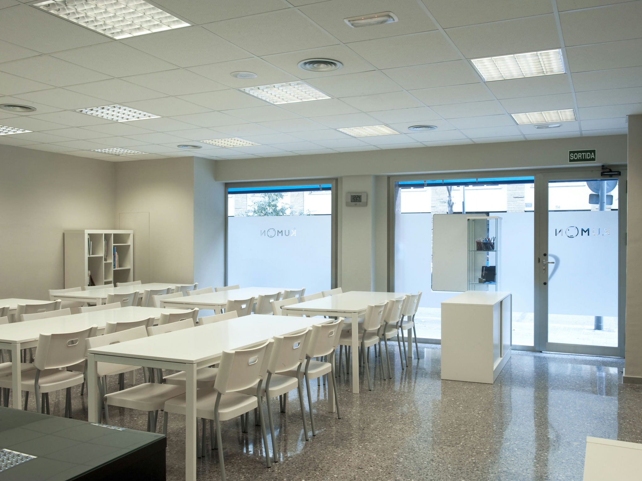 Una aula moderna y luminosa y espaciosa que presenta mesas y sillas blancas dispuestas en filas, grandes ventanas que permiten la entrada de luz natural, paredes de color neutro, suelo pulido, una pequeña estantería y un escaparate minimalista cerca de la entrada.
